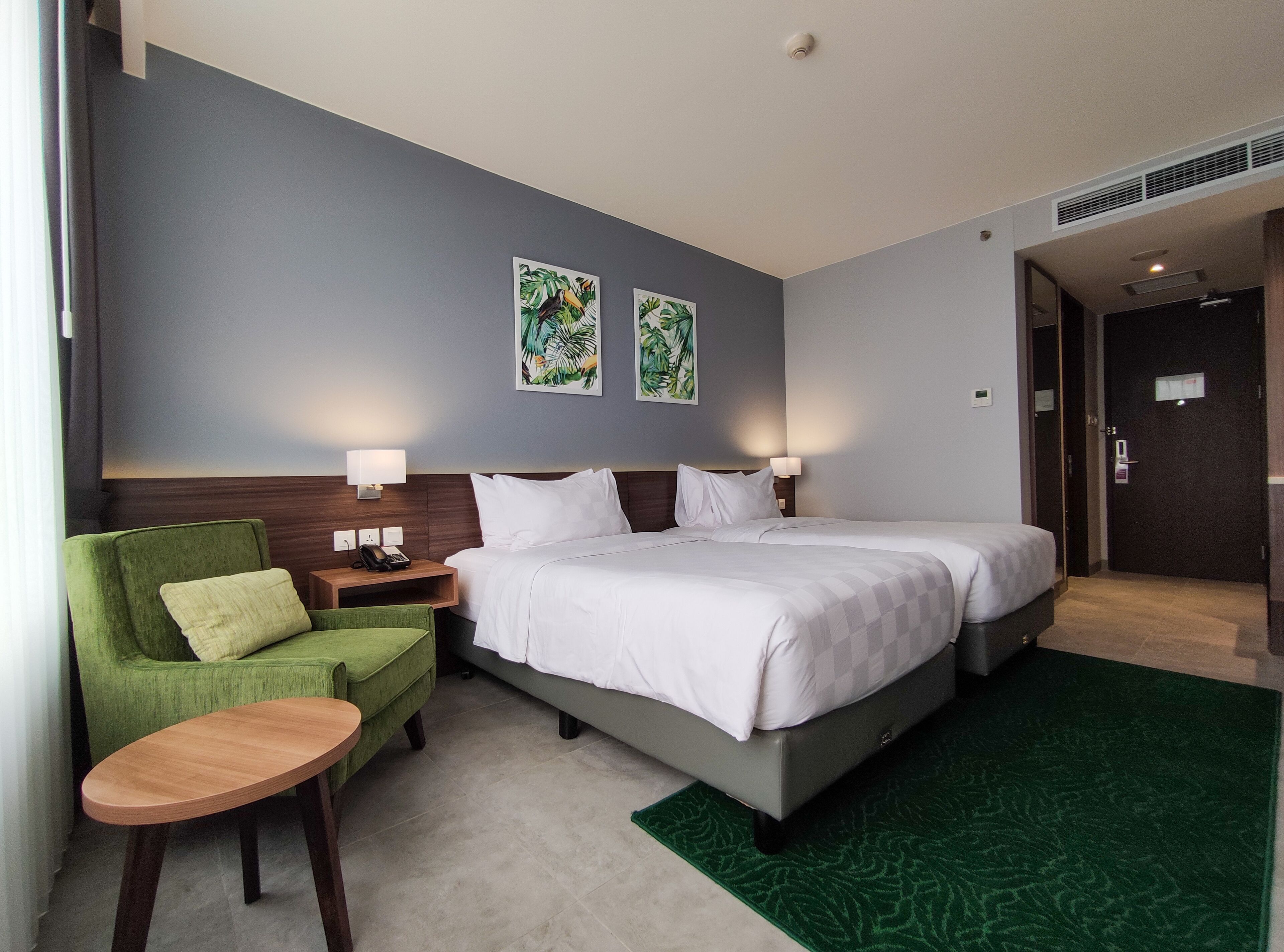 Une chambre d’hôtel avec un lit, un fauteuil vert, une petite table en bois et deux lampes fixées au mur.