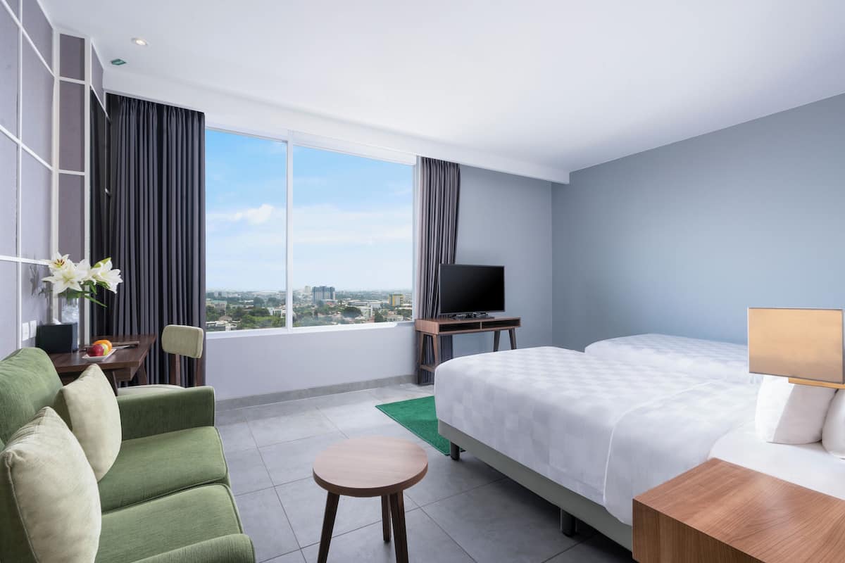 A hotel room with a bed, a green sofa, a small table, a TV, and a large window offering a city view.