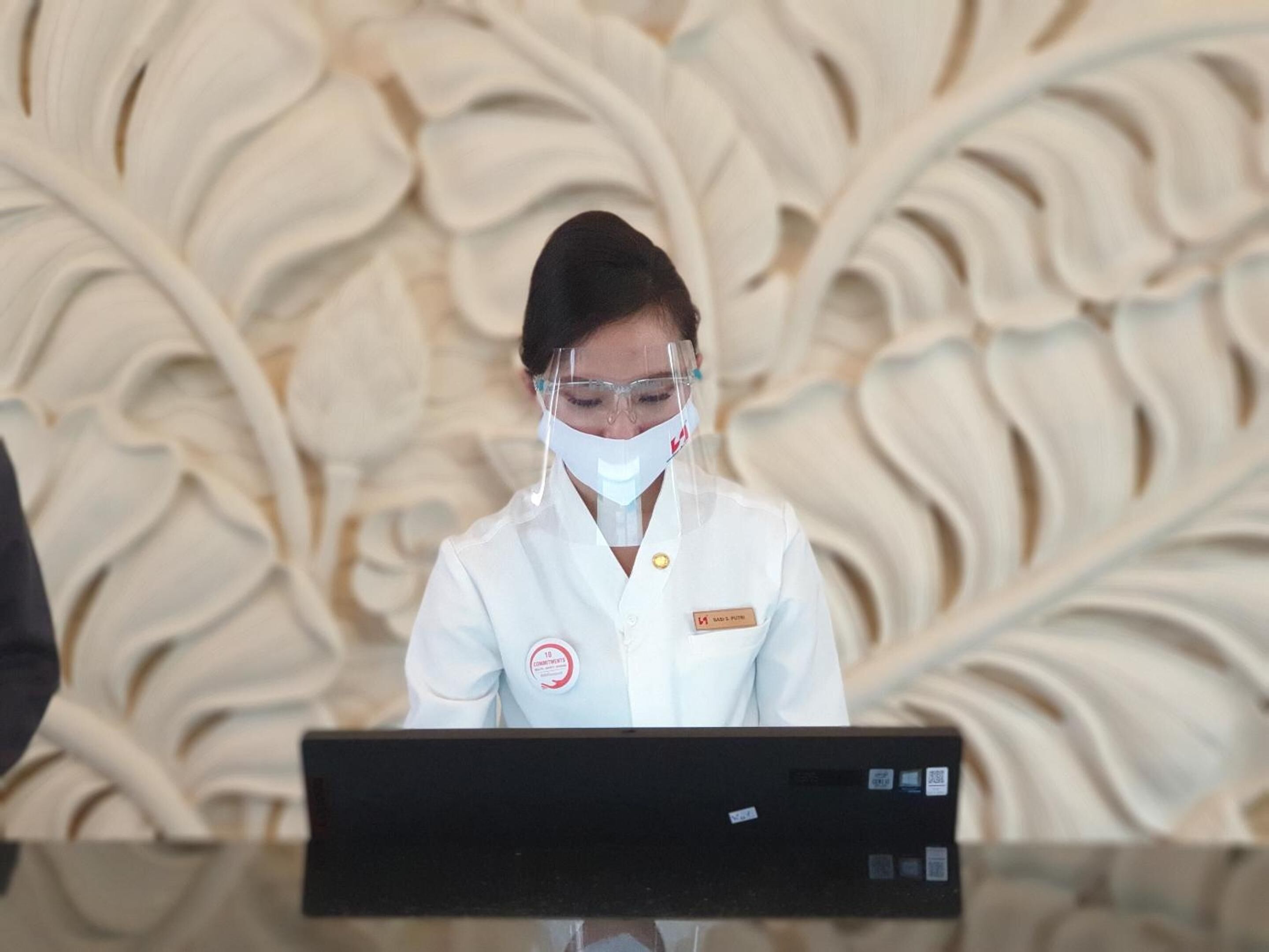 A person in a white uniform with a name tag and a face shield, standing in front of a black counter.