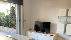 A modern living room with a flat-screen TV on a white cabinet, a grey sofa, and a glass door leading to a balcony with plants. - Ground floor Aqua Nature La Mata Costa Blanca (La Mata)