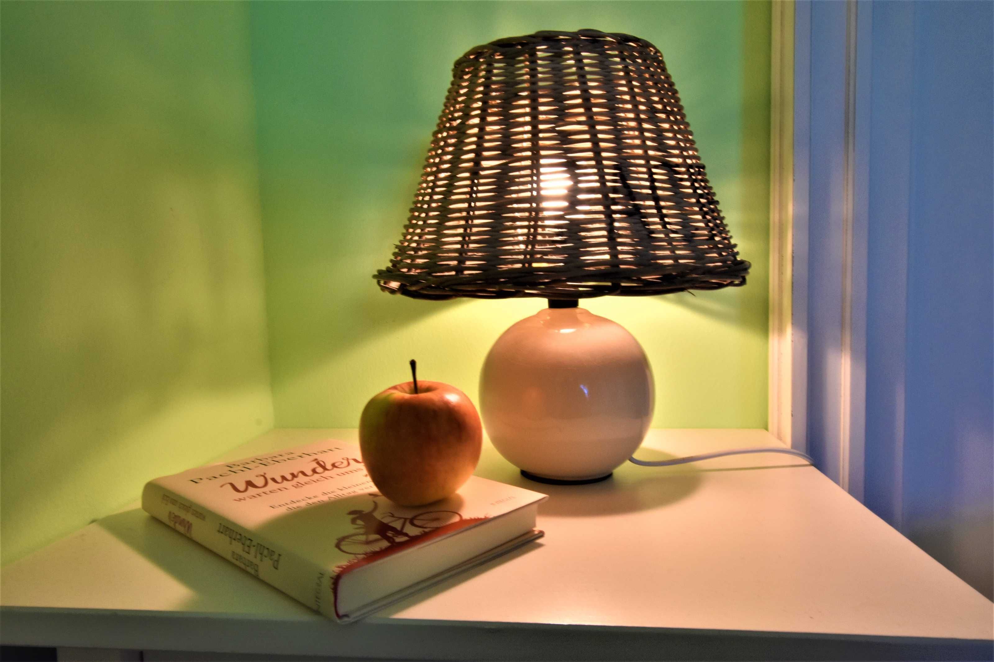 A lamp with a woven shade, an apple, and a book on a white surface.