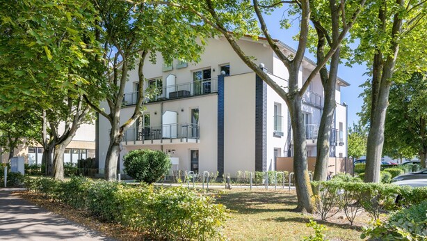 Un immeuble résidentiel à plusieurs étages, doté de balcons, d’une allée pavée et entouré de verdure.