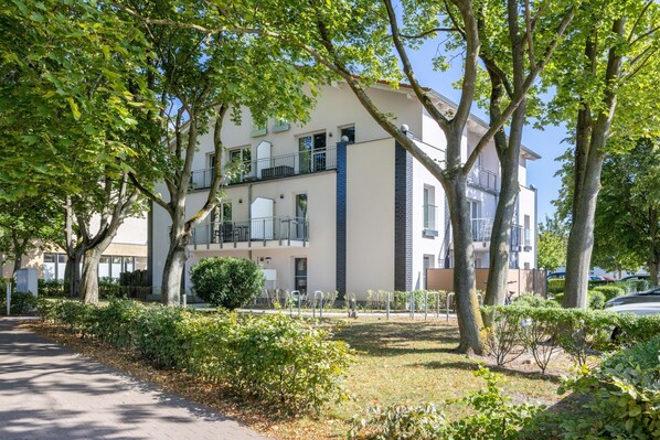 Un edificio residencial de varios pisos con balcones, un camino de entrada pavimentado y vegetación circundante.