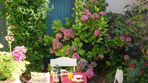 A red table with a basket of bread, a coffee pot, and a glass of orange juice, set against a backdrop of lush greenery and pink hydrangea flowers. - Historic 18th Century Home with Modern Comforts in Medieval Village (Montmirail)