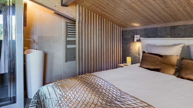 Une chambre avec un plafond en bois, un lit recouvert d’une couvre-lit à motifs, une table de chevet avec une bougie et un luminaire fixé au mur.