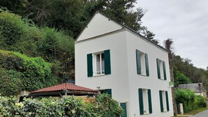 A two-story building with green shutters, a red-tiled roof, and surrounded by dense greenery.