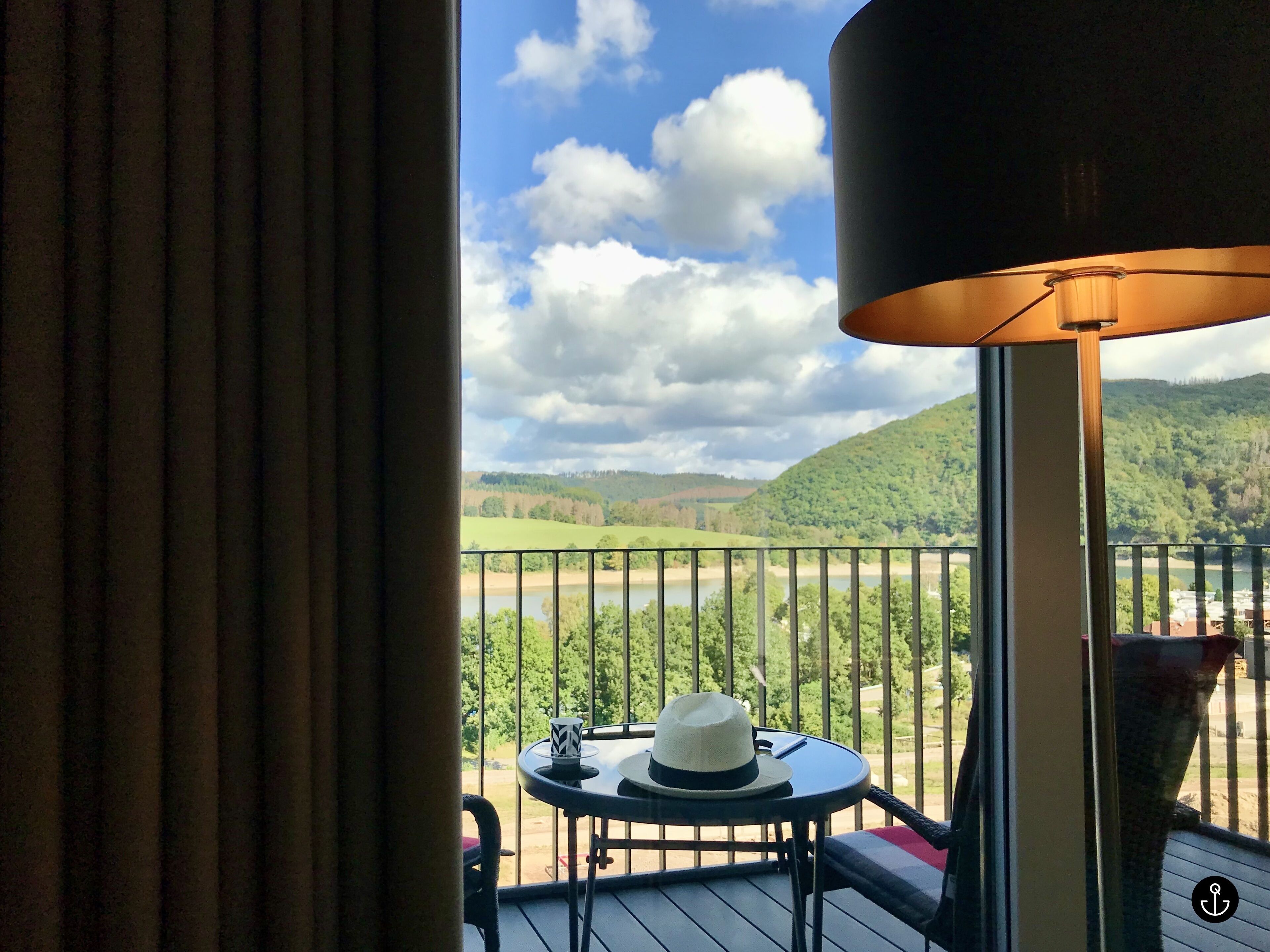 A balcony with a view of a river and hills, a table with a hat, and a lamp.
