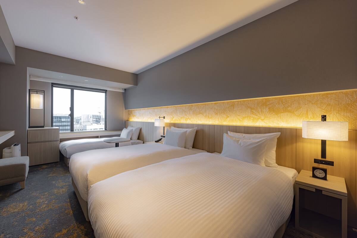 A hotel room with two beds, a desk, a chair, a nightstand with a lamp, and a large window offering a city view.