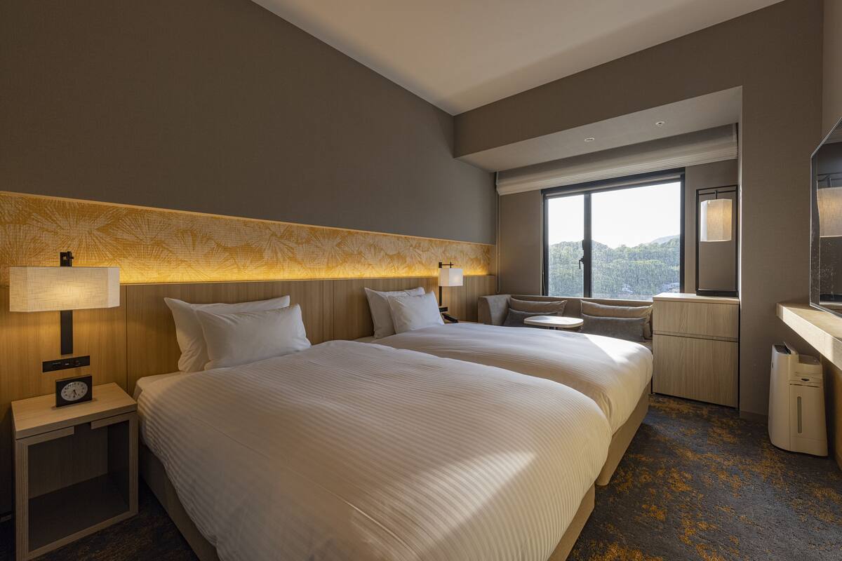 A hotel room with two beds, a desk, a chair, a mirror, and a view of the outdoors.