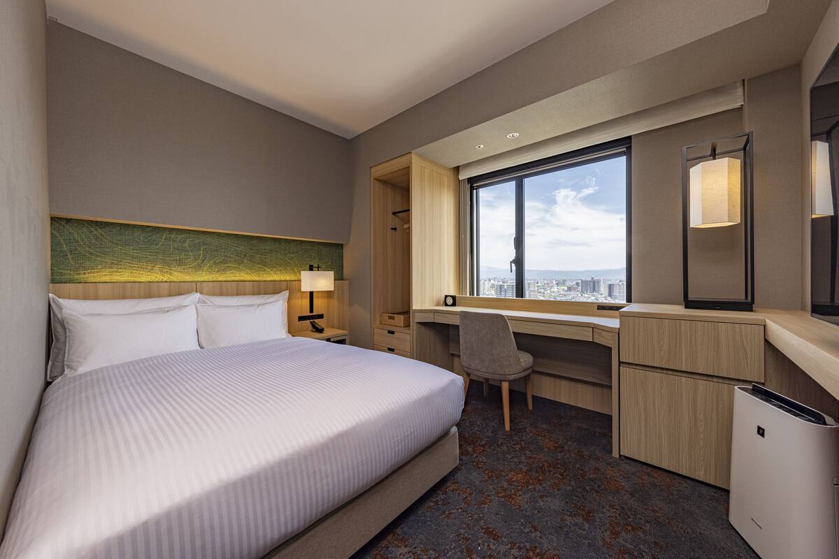 A hotel room with a large bed, a desk, a chair, and a view of the city through a large window.