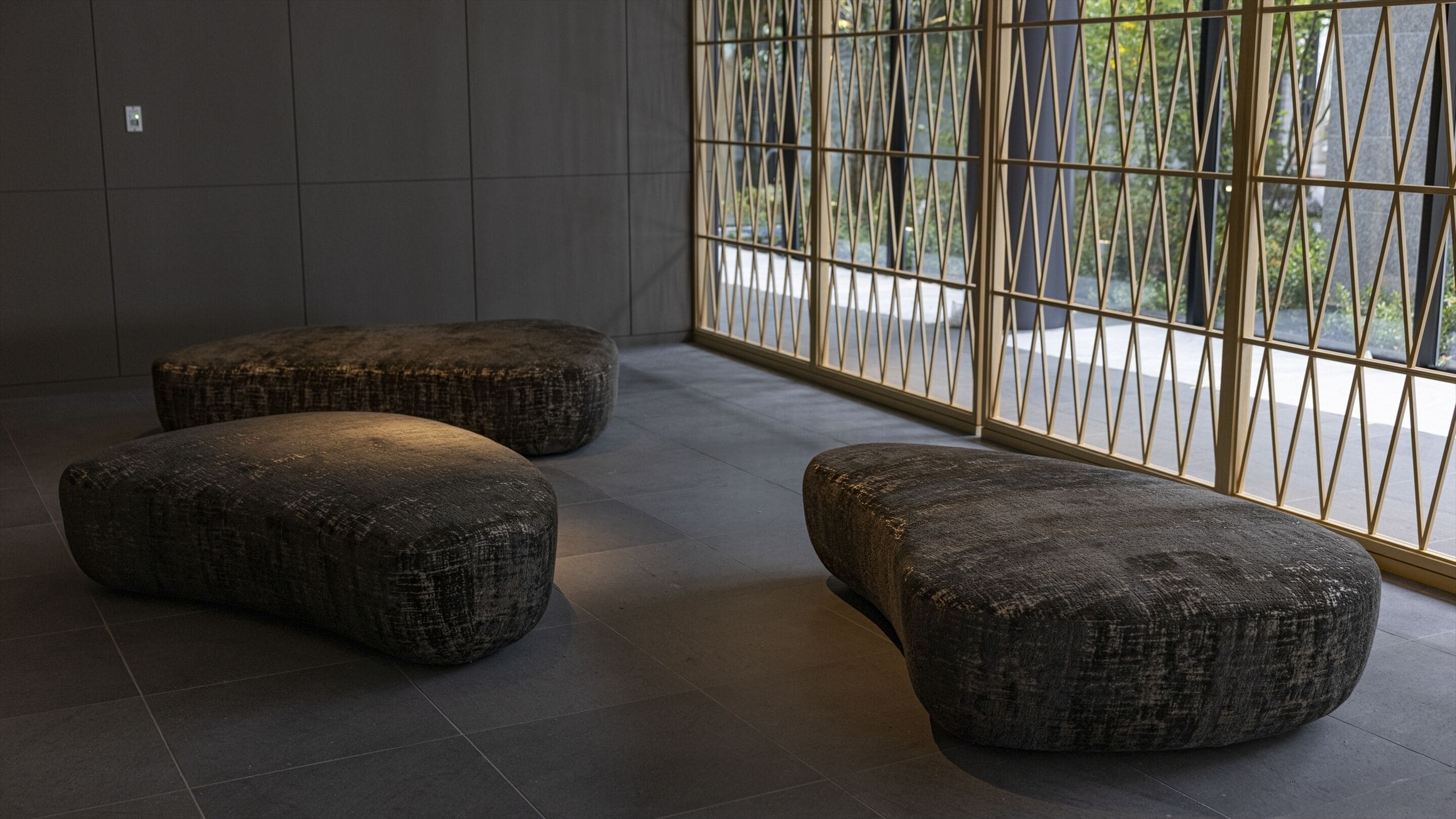 Een modern interieur met drie donkere, gestructureerde poefs en een groot raam met een geometrisch metalen frame.