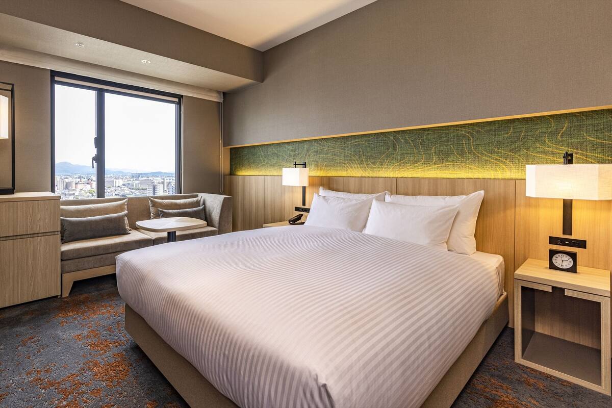 A hotel room with a large bed, a desk, a sofa, and a view of the city.