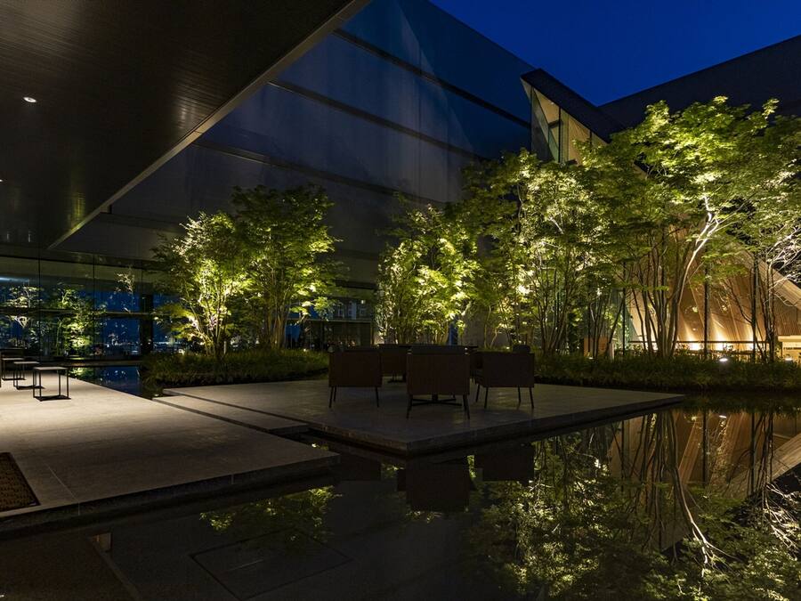 Un espacio exterior moderno con asientos, un estanque reflectante y árboles iluminados con luz suave.