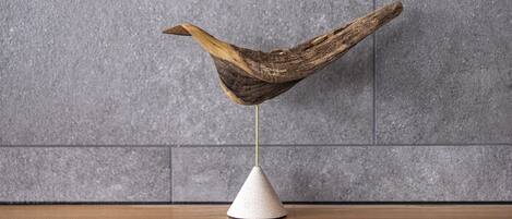 Une sculpture en bois, posée sur un piédestal blanc, se détache sur un mur recouvert de carreaux gris.