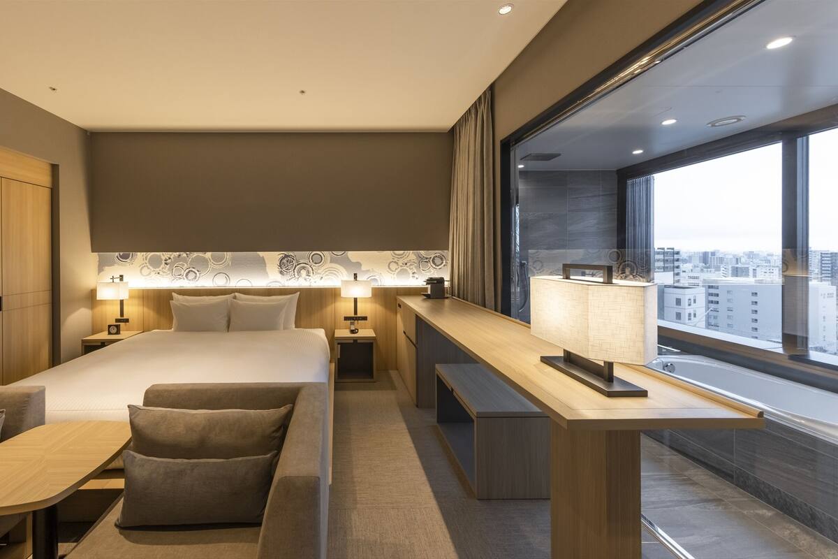A modern hotel room with a large bed, bedside tables, a desk, and a view of the city.