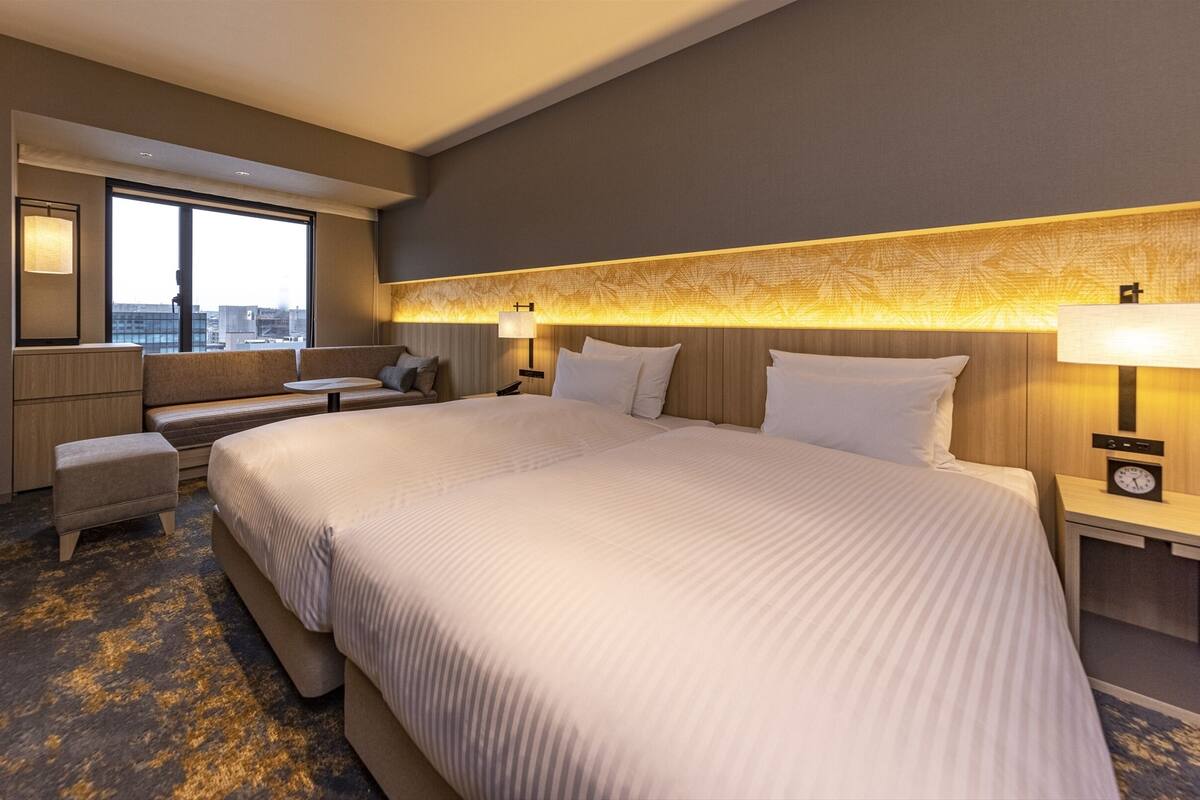 A hotel room with a large bed, a sofa, a desk, and a window with a city view.