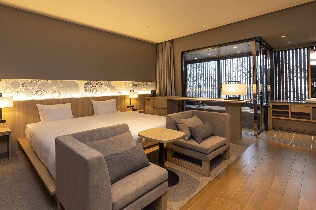 A modern hotel room with a large bed, two armchairs, a small table, and a bathroom visible through a glass partition.