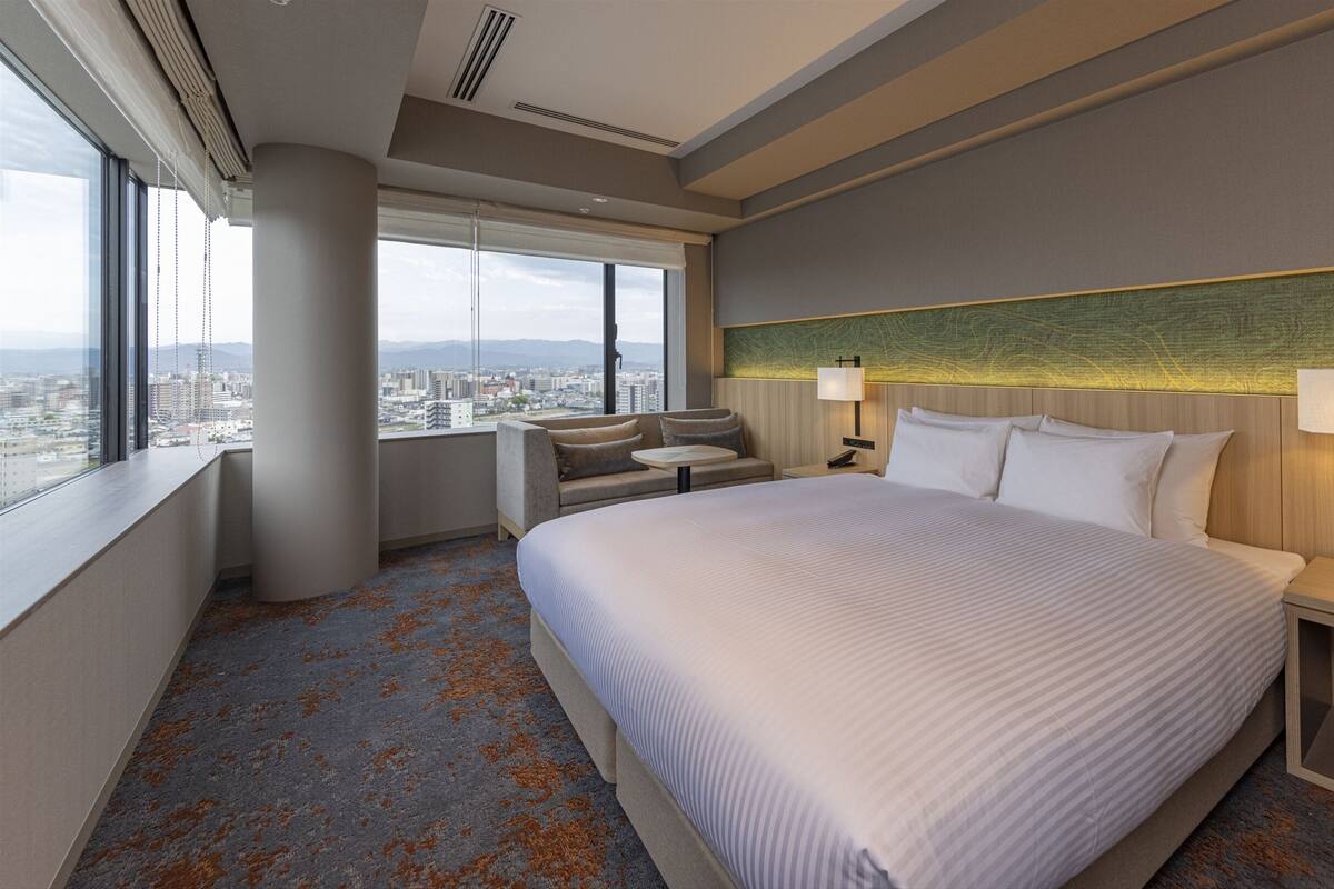 A hotel room with a large bed, two bedside tables with lamps, a sofa, and a view of the city through large windows.