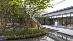 A modern building with a glass facade, a reflective water feature, and a landscaped area with trees and plants. - THE BLOSSOM KUMAMOTO (Kumamoto)