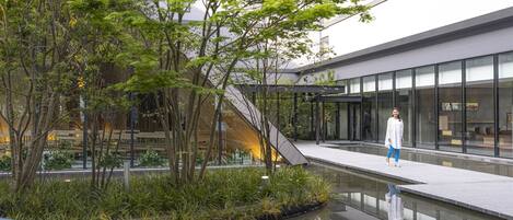 Un bâtiment moderne doté d’une façade en verre, d’un élément décoratif aquatique réfléchissant et d’un espace paysager agrémenté d’arbres et de plantes.