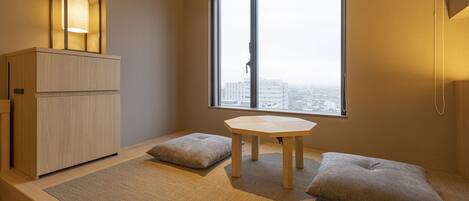 Habitación minimalista con un ventanal grande, una mesa de madera, dos cojines y un mueble con una lámpara.