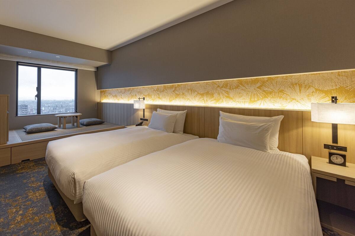 A hotel room with two beds, a small table, a chair, a window with a city view, and a nightstand with a lamp and clock.