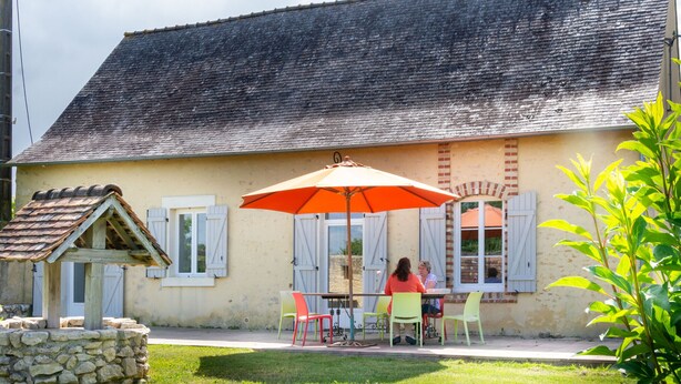 A building with a sloped roof and a stone foundation. There is a table with chairs and an umbrella outside.