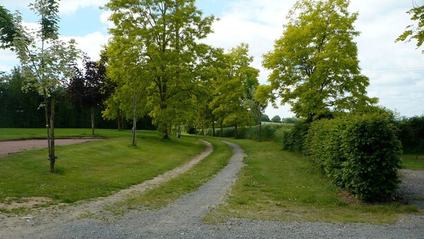Un sentier de gravier serpente à travers un parc, bordé d’arbres et d’une haie.