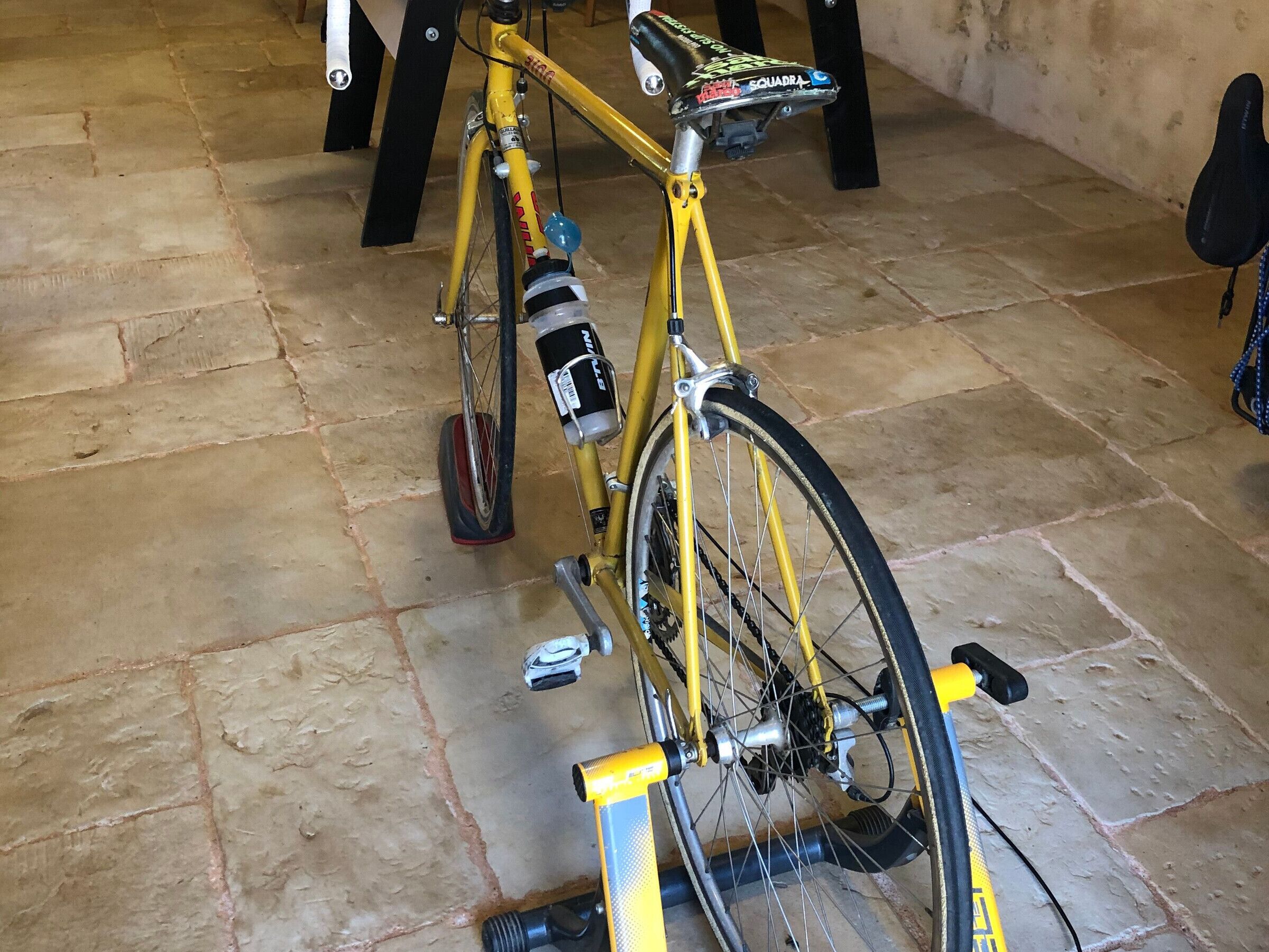 Un vélo jaune, muni d’un porte-bidon et d’une sacoche fixée au siège, est garé à l’intérieur, sur un sol carrelé.