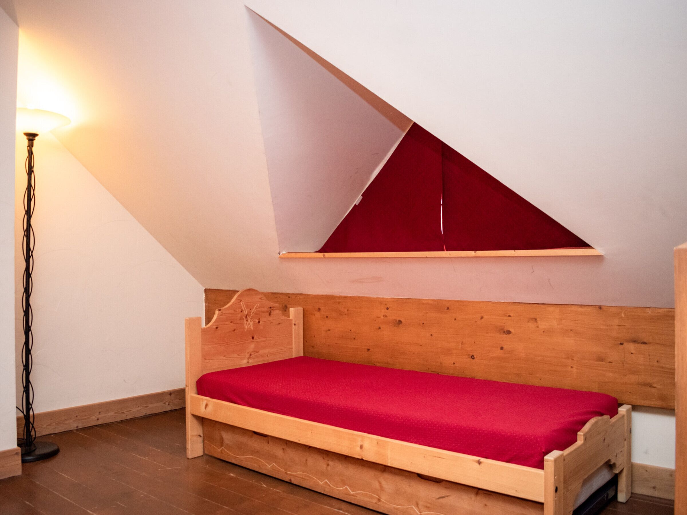 Un petit lit en bois dans une pièce dont le plafond est en pente et qui possède une fenêtre triangulaire rouge.