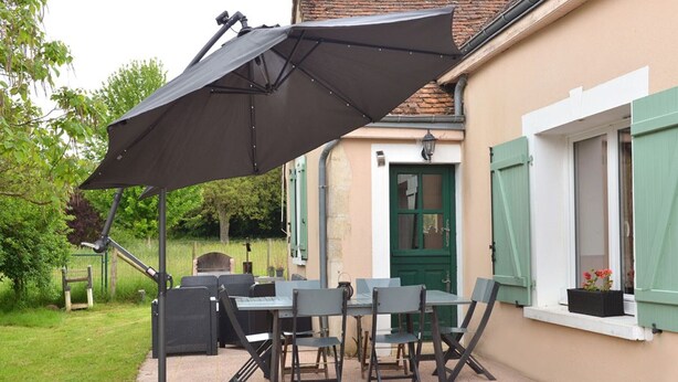 An outdoor patio with a table and chairs, a large umbrella, and a house with green shutters.
