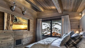 Ein gemütliches Schlafzimmer mit einem großen Bett, einem aufgehängten Hirschgeweih, einer hölzernen Kommode und einem Fenster mit Blick auf einen verschneiten Berg.