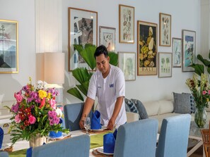 A person setting a table with a floral centerpiece, a framed picture of Bali, and a sofa in the background. - Villa Lulito (Seminyak)