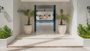 Une entrée moderne donnant sur la piscine, avec une façade blanche, deux palmiers en pot et un cadre de porte bleu.