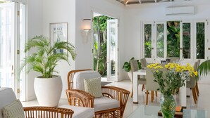 Une pièce spacieuse comprenant un coin repas, des meubles en osier, une grande plante en pot et une table en verre sur laquelle sont posés des livres.