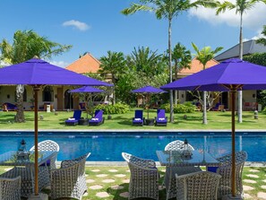 A poolside area with lounge chairs and umbrellas, a clear blue pool, and a building with a tiled roof in the background. - Sayang d’Amour (Seminyak)