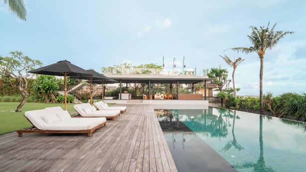 A poolside area with lounge chairs and umbrellas, a wooden deck, and a view of a building with a covered seating area. - Arnalaya Beach House (Canggu)