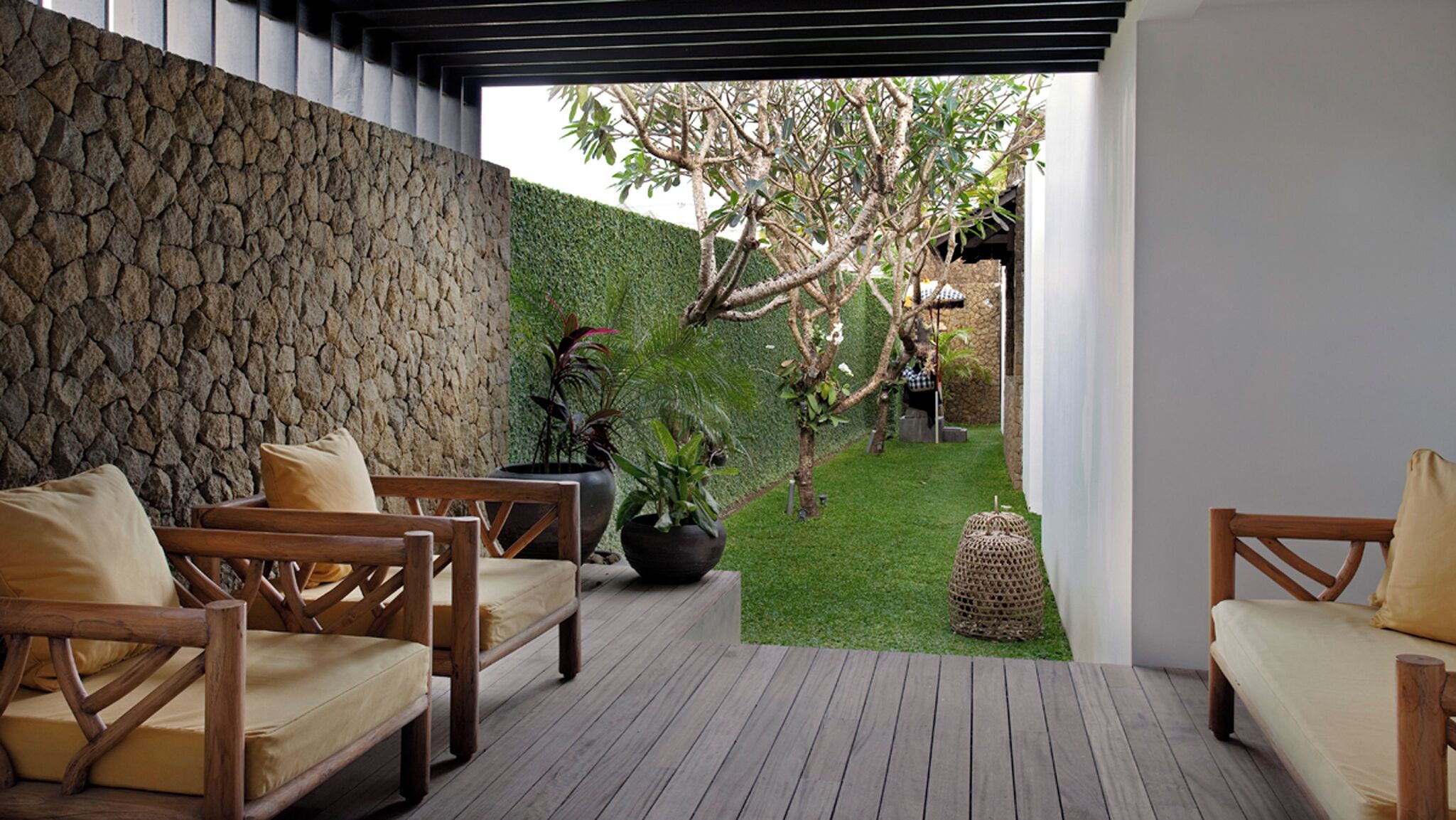 A wooden deck with outdoor seating, a stone wall, and a view of a garden with a tree and a hedge.