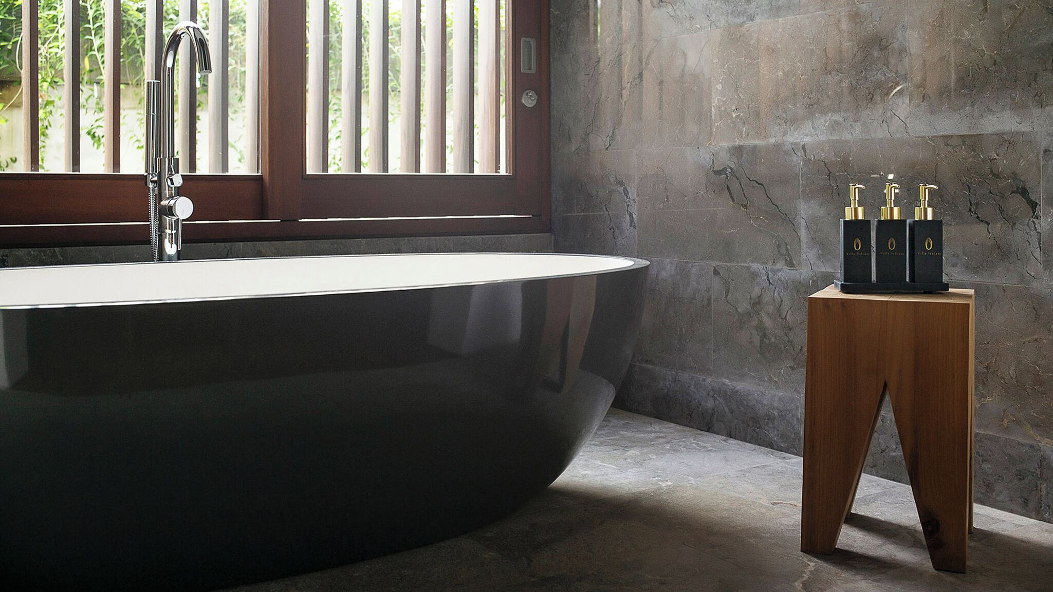 A modern bathroom with a freestanding bathtub, a wooden stool with bottles, and a window with vertical blinds.