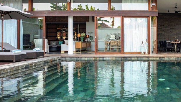 A modern pool area with lounge chairs and a bar area visible through glass doors. - Villa Indrani (Canggu)