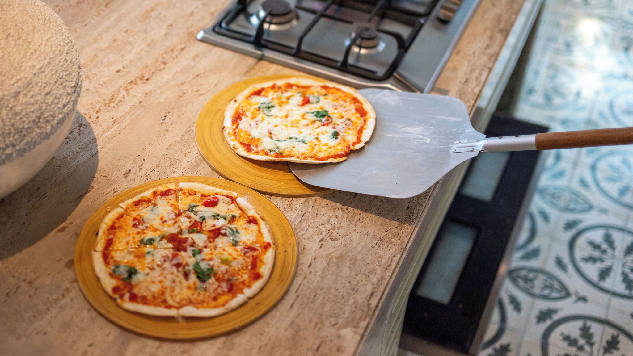 Dos pizzas sobre platos de madera en una encimera de cocina, junto a una pala para pizza.