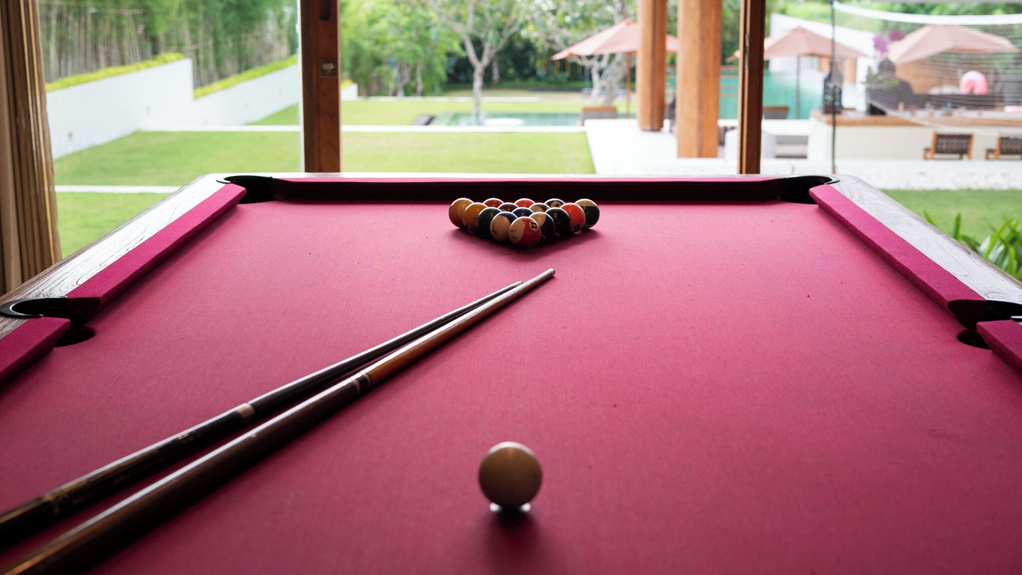 A pool table with a cue stick and billiard balls, set against a view of a garden and outdoor seating area.