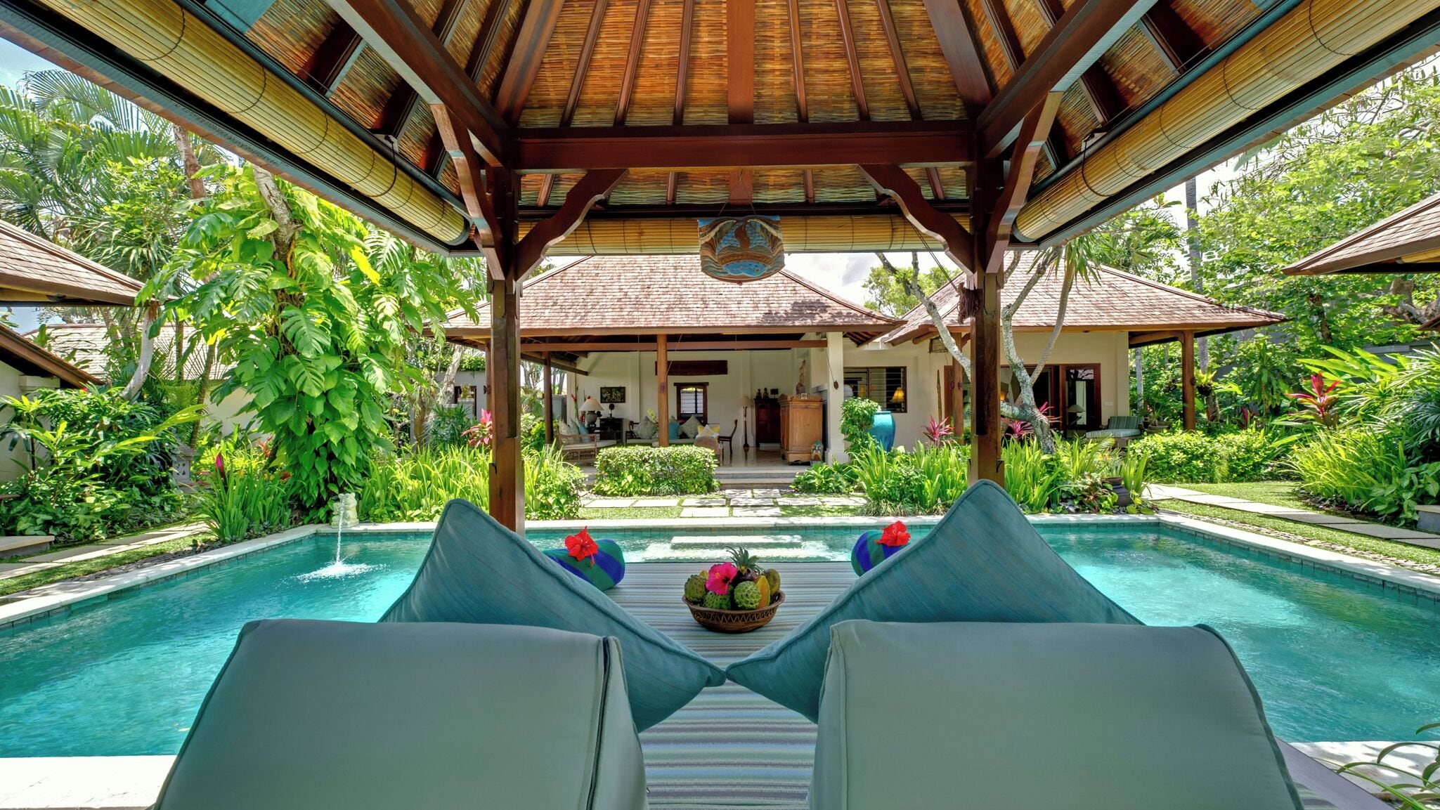 Un área de piscina con sillones reclinables, un espacio de estar cubierto y abundante vegetación tropical.