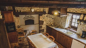 A rustic kitchen with a stone wall, wooden cabinets, a tiled backsplash, a built-in oven, a sink, and a dining table with chairs. - Charming Riverside Cottage with Fishing & Wifi, Near Historic Sites (Rouez)