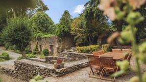 A rustic stone patio with wooden furniture, surrounded by lush greenery and a stone building. - Charming Riverside Cottage with Fishing & Wifi, Near Historic Sites (Rouez)