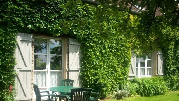 A patio with a round table and chairs, surrounded by a stone border, under a covered area with white shutters and lush greenery. - Charming Farmhouse with Fireplace, Garden, and Modern Amenities in Joué-l'Abbé (Joué-l'Abbé)