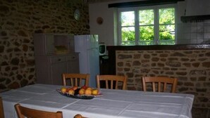 A dining area with a stone wall, a window with a view of greenery, a table with a white tablecloth, and wooden chairs. - Charming Farmhouse with Fireplace, Garden, and Modern Amenities in Joué-l'Abbé (Joué-l'Abbé)