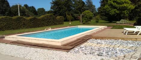A rectangular swimming pool with a tiled deck surrounding it, set in a landscaped garden with trees and shrubs.