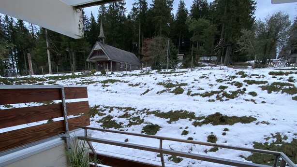 Ein Balkon mit Glastisch und Stühlen, einem Geländer und Blick auf eine verschneite Landschaft mit Bäumen und einem Gebäude im Hintergrund.