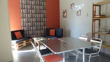 A modern library with a long bookshelf, a black sofa with orange cushions, a glass-top table, and metal chairs.